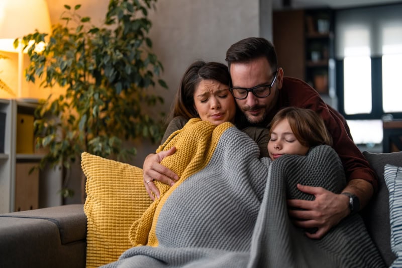 Family of three huddled under a blanket trying to keep warm.