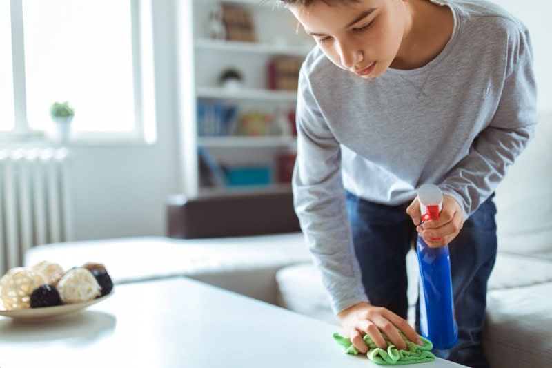 Child cleaning the top of a table with a spray and rag.